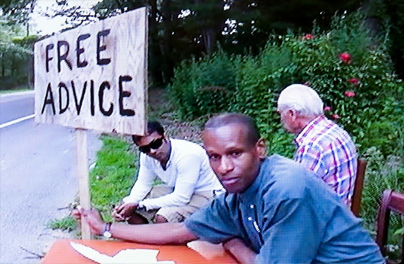 Three people sitting at a table on the side of a road, one person holding a 'Free Advice' sign