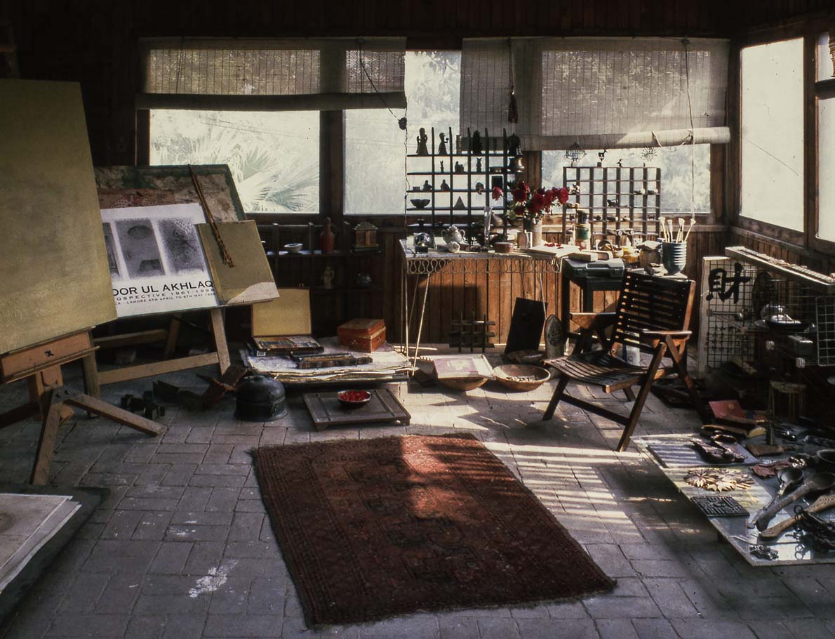 Artist Zahoor ul Akhlaq's studio, with media on easels, ceramics and brushes on a table in the back, various objects on the ground, and a small rug and chair